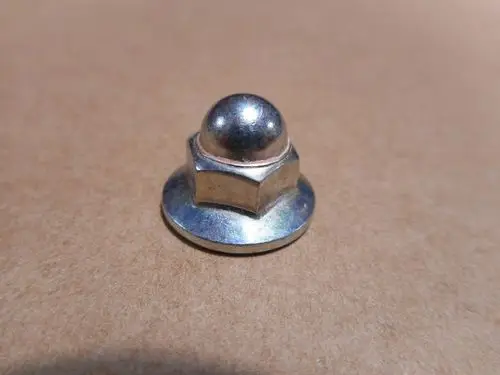 Mutter aus Metall mit einer gewölbten Oberseite und einer sechseckigen Basis, auf einem beige Hintergrund platziert. Herstellerangabe: Remeha.